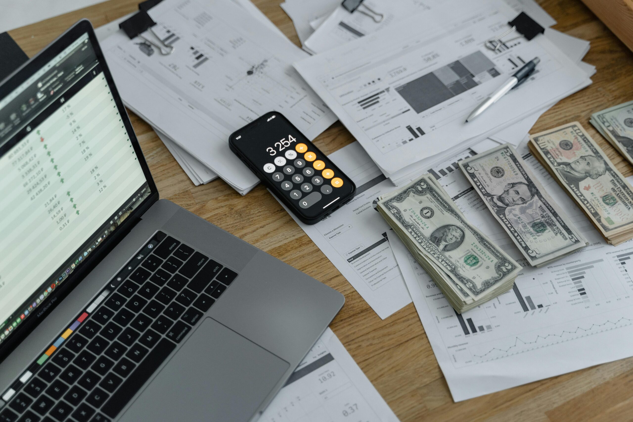 Workspace with laptop displaying financial spreadsheets, a calculator app on a smartphone, and stacks of U.S. currency alongside printed financial documents.