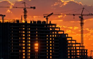 Image of a silhouette of a multi-story building under construction at dusk with two construction cranes in the background. This image is used to illustrate a blog post about construction debt collection and mechanic's liens.