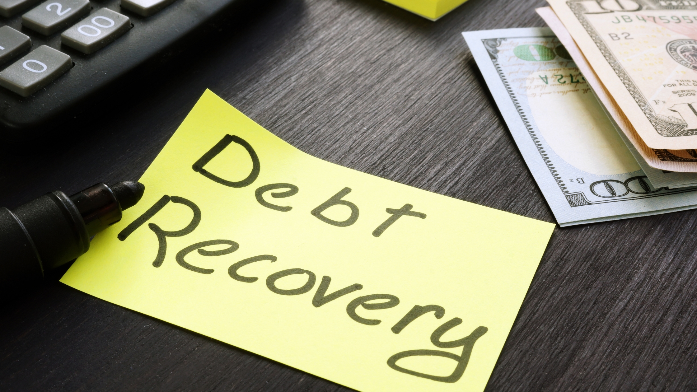 Sticky note with "Debt Recovery" written on it placed on a desk next to a calculator.