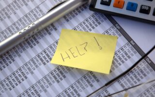 Close-up of financial report with yellow sticky note showing “Help!”. Track down unpaid invoices with The Baker Group's debt collection services.