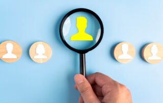 Hidden Cost of DIY Debt Collection: Close-up of hand holding magnifying glass inspecting blue employee icon on computer screen.