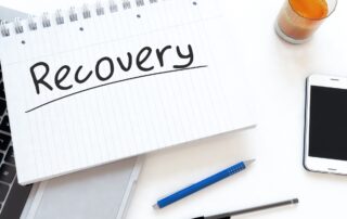 International Debt Collection: A notebook page with "Recovery" written on it, accompanied by a pen, phone, and laptop, representing the tools used in debt collection.